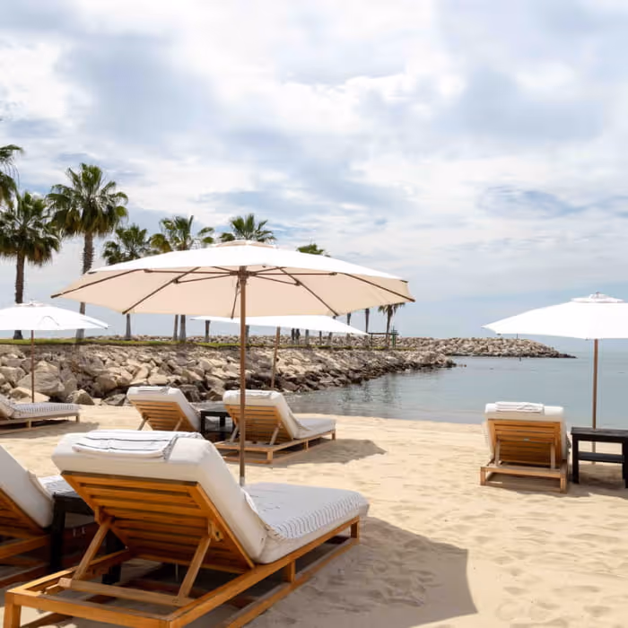 Sandy beach resort with wooden lounge chairs, white cushions, and large umbrellas overlooking calm ocean waters lined