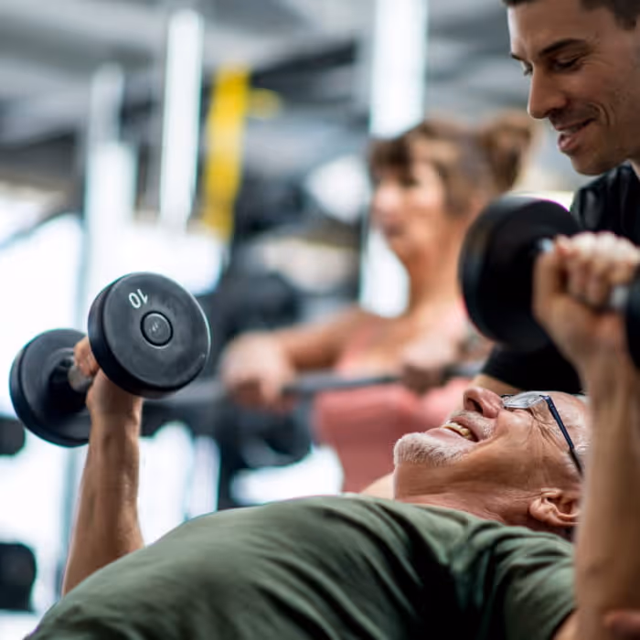 Older man wearing glasses smiles while lifting 10-pound dumbbells on a bench press as a personal trainer spots him
