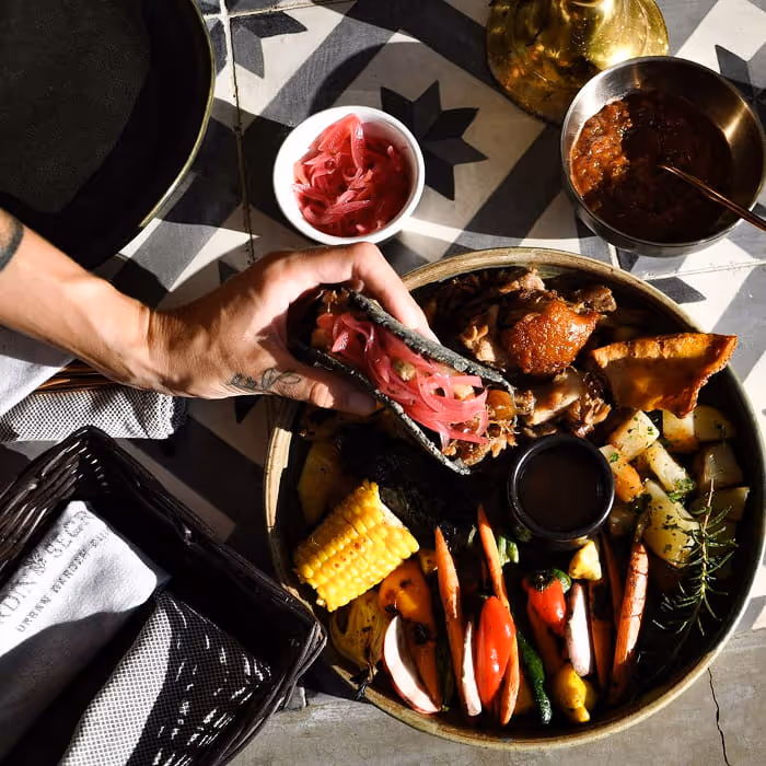 Hands assembling a blue corn taco with pickled red onions from a platter of roasted meats, grilled vegetables, and corn