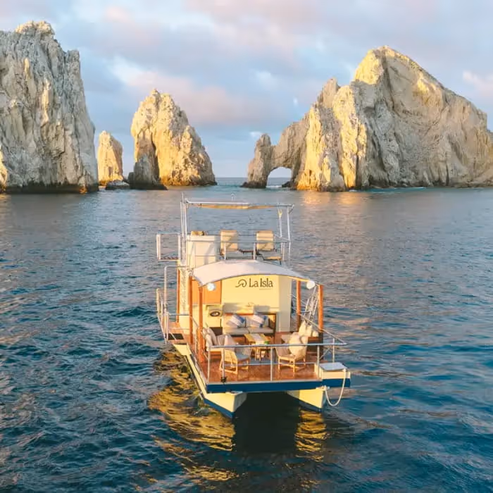 La Isla boat floating near the iconic El Arco rock formation in Cabo San Lucas during a golden sunset over the sea.