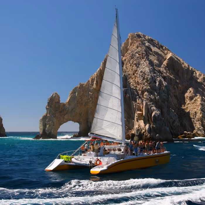 A catamaran sailboat full of tourists cruises past the iconic El Arco rock formation in Cabo San Lucas, Mexico.