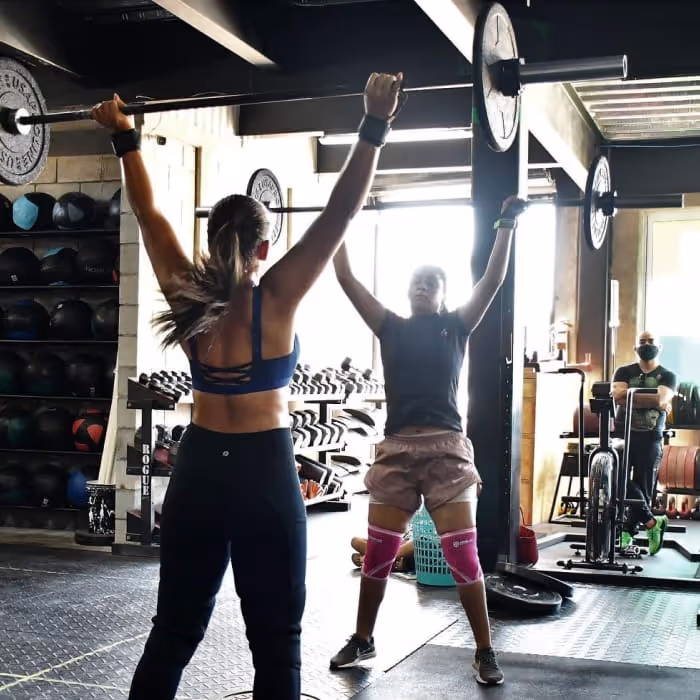 Two athletes performing overhead barbell presses together in a well-equipped CrossFit gym with Rogue equipment