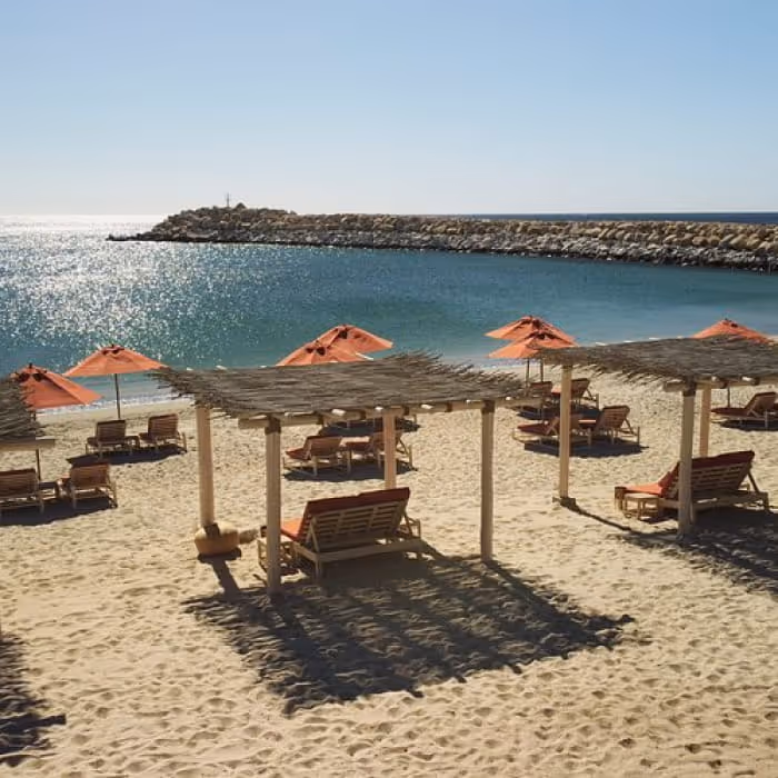 Sandy resort beach with thatched palapas, orange lounge chairs, and umbrellas overlooking a calm turquoise cove