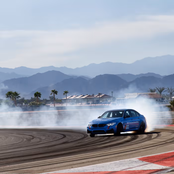 Blue BMW M3 drifting on a racetrack with tire smoke billowing, mountains visible in the background