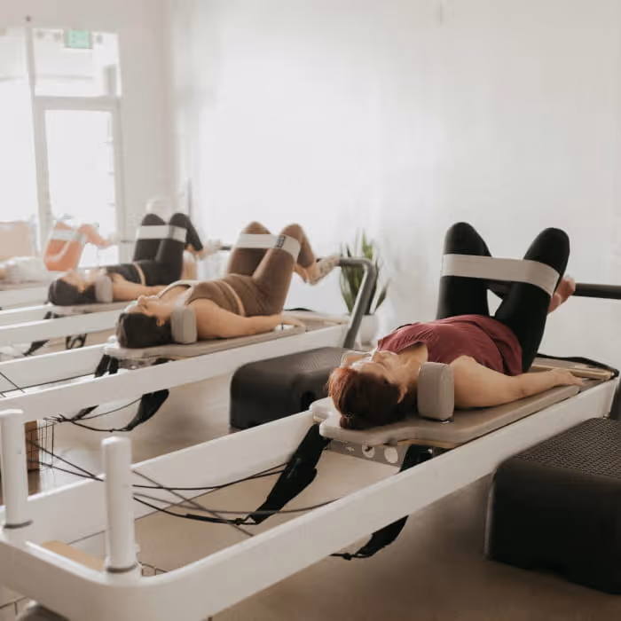 Three women lying on Pilates reformer machines performing leg exercises in a bright, minimalist fitness studio