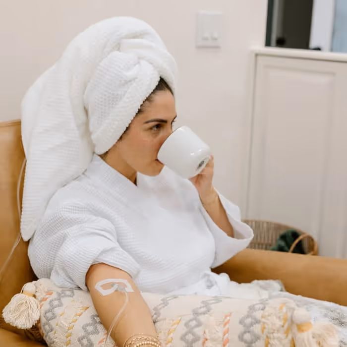 Woman in a white robe and towel-wrapped hair sipping from a mug while receiving an IV drip therapy treatment