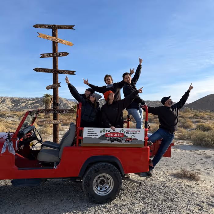 Group of five excited tourists posing on a Red Jeep Tours vehicle in a desert landscape near directional road signs.