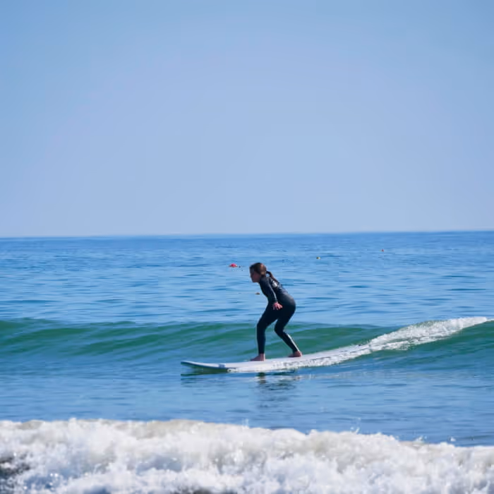 A surfer in a black wetsuit riding a small wave on a longboard in calm blue ocean water under a clear sky.