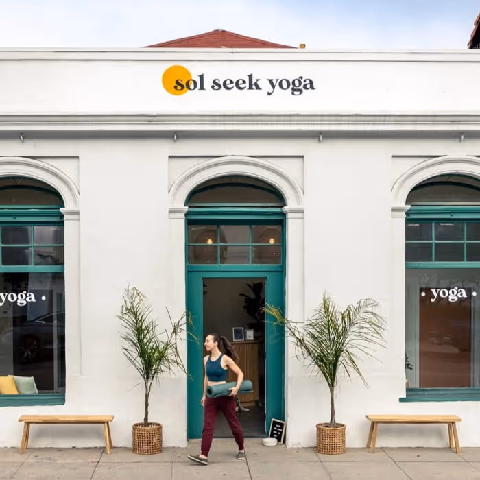 A woman carrying a yoga mat walks past the entrance of Sol Seek Yoga studio, a white building with teal-framed windows.