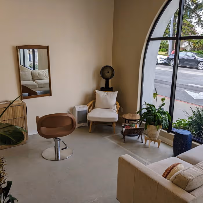 A cozy hair salon interior with a styling chair, mirror, large arched window, indoor plants, and neutral-toned lounge
