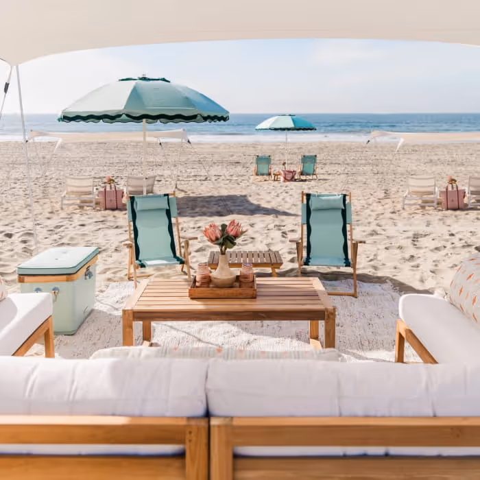 A luxurious beach picnic setup with teak furniture, mint-green chairs, umbrellas, and ocean views on a sandy shoreline.