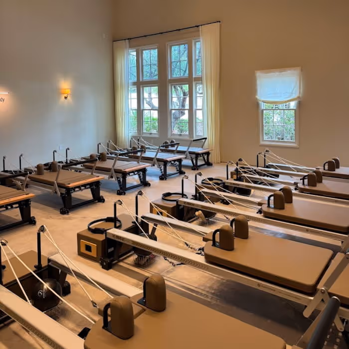 A Pilates reformer studio with multiple machines arranged in rows inside a bright room with large windows and warm lighting.