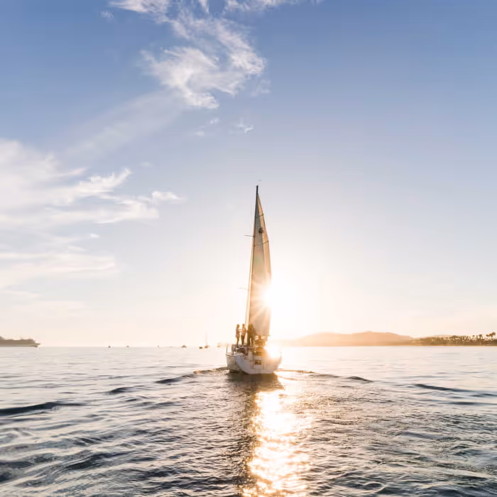 A sailboat glides across calm ocean waters at sunset with sunlight reflecting off the sea and distant hills on the horizon.