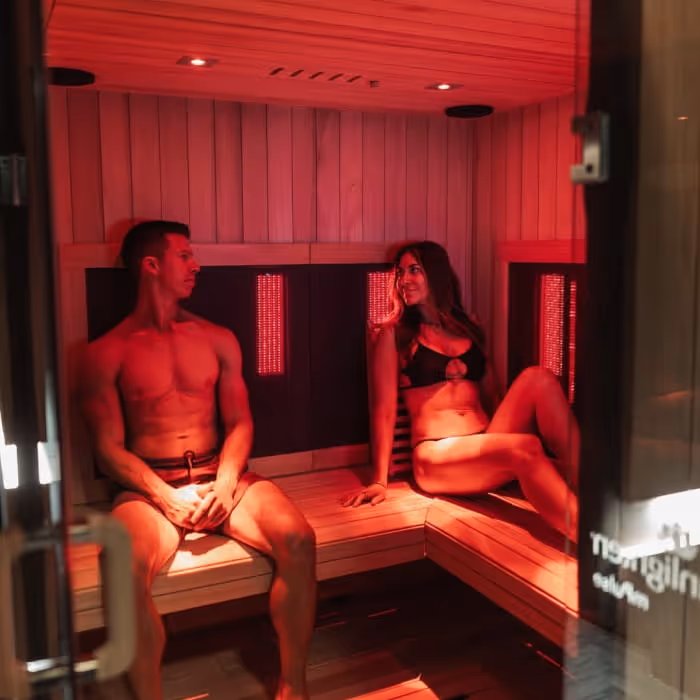 A couple relaxing inside an infrared sauna with red lighting, sitting on wooden benches in swimwear and smiling at each