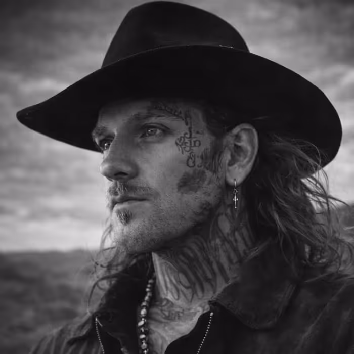 Black and white portrait of a man with face and neck tattoos wearing a wide-brimmed hat and a cross earring.