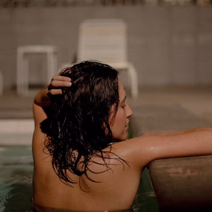 Woman with wet dark hair pushes her hair back while relaxing at the edge of a swimming pool at night.