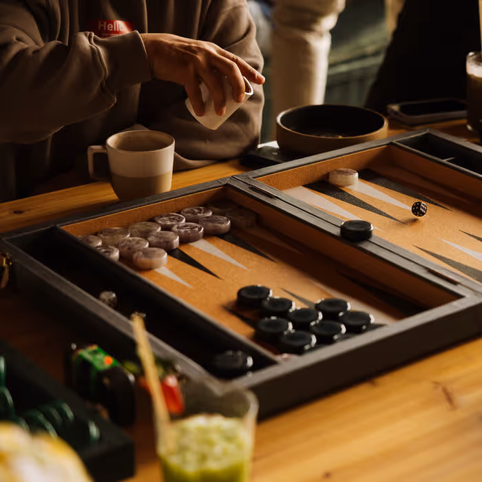 A person reaches for a coffee cup beside a backgammon board with marble and black game pieces on a wooden table.