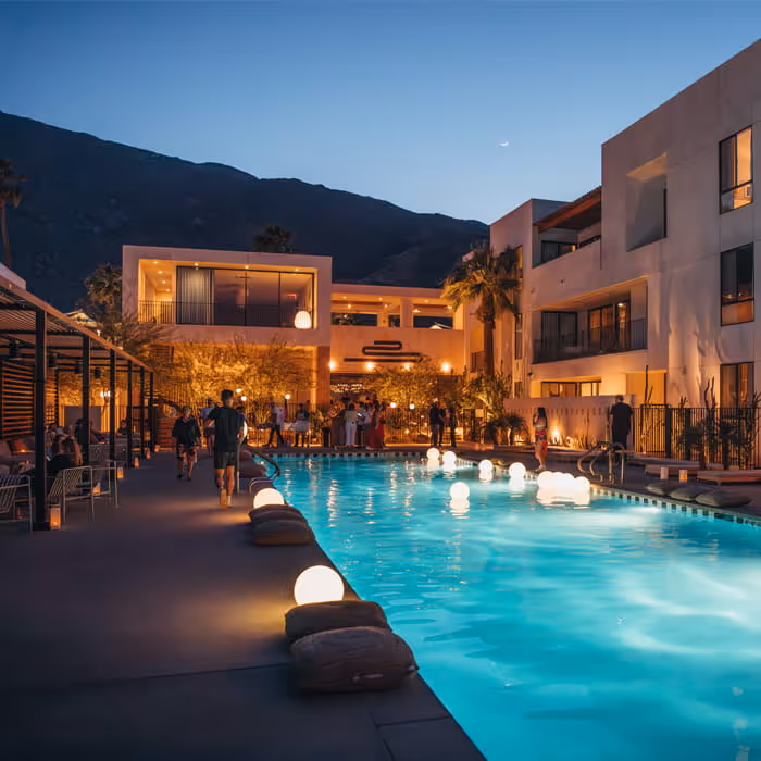 A luxury desert hotel pool at twilight with glowing orbs floating in the water and mountains in the background.