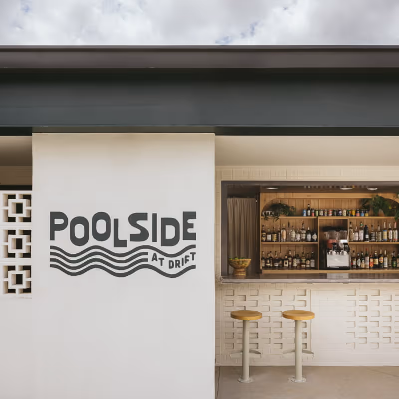 Poolside at Drift outdoor bar featuring wooden shelves stocked with bottles, two yellow stools, and white decorative brick