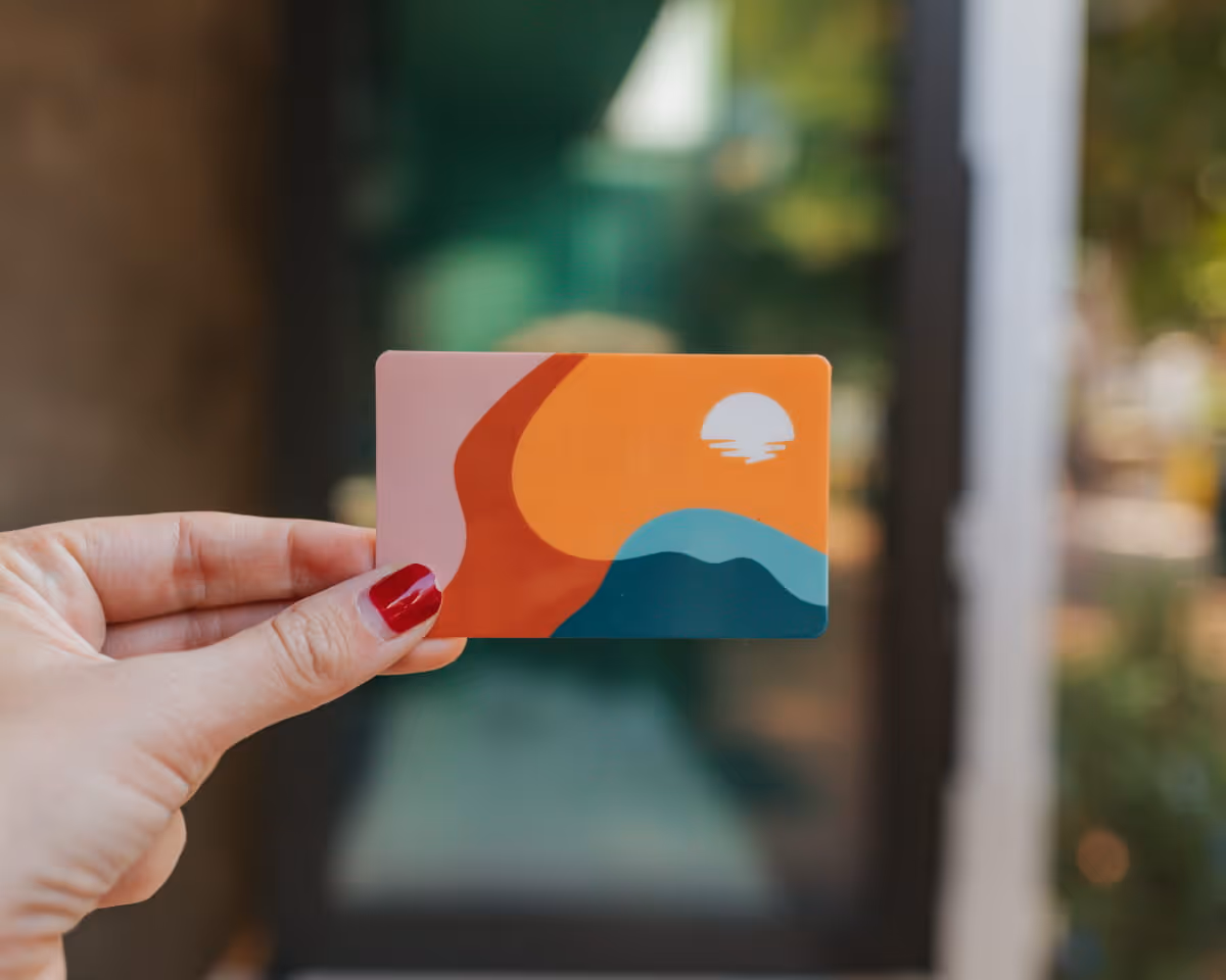 A hand with red nail polish holds a colorful gift card featuring a retro sunset design with orange, pink, and teal tones.