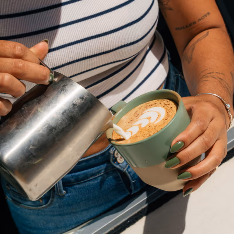 A barista with green nails pours steamed milk from a metal pitcher into a sage green cup, creating latte art on the coffee.