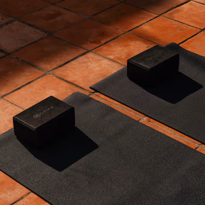 Two black Gaiam yoga blocks placed on dark yoga mats arranged on warm terracotta tile flooring