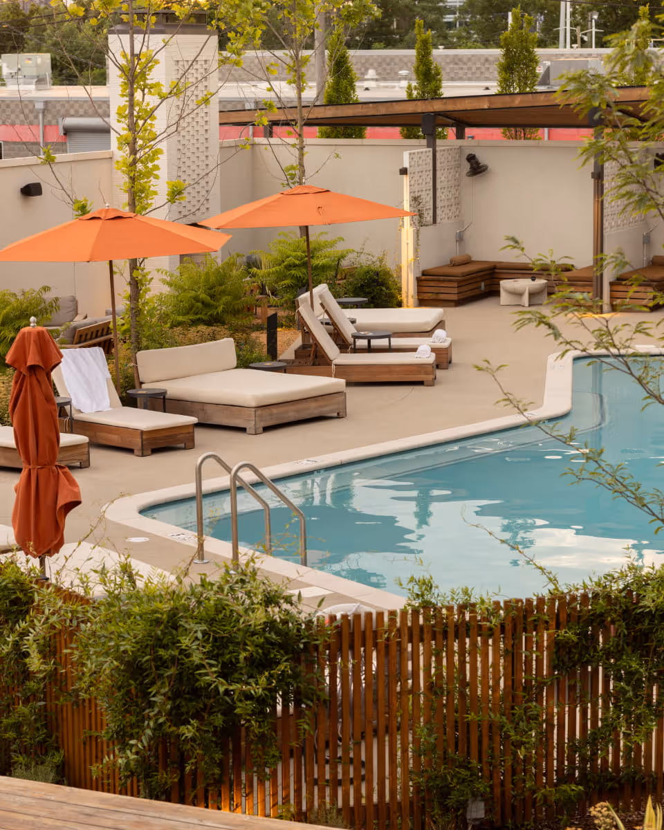 Hotel pool area with cream cushioned lounge chairs, orange umbrellas, lush greenery, and a wooden fence surrounding the deck.
