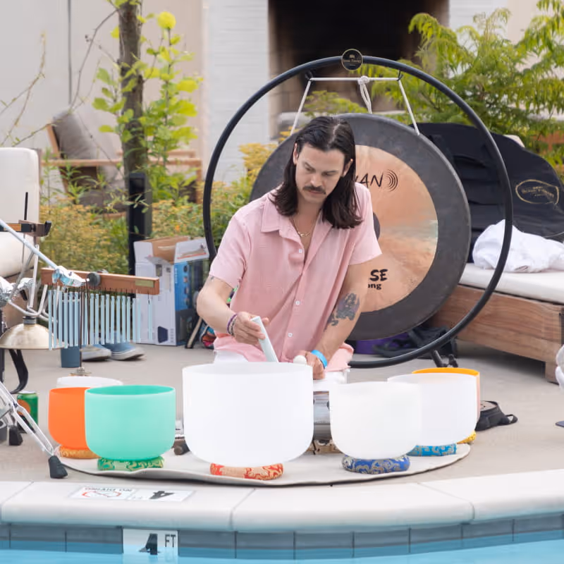 A sound healer in a pink shirt plays crystal singing bowls beside a pool, with a large gong positioned behind him.