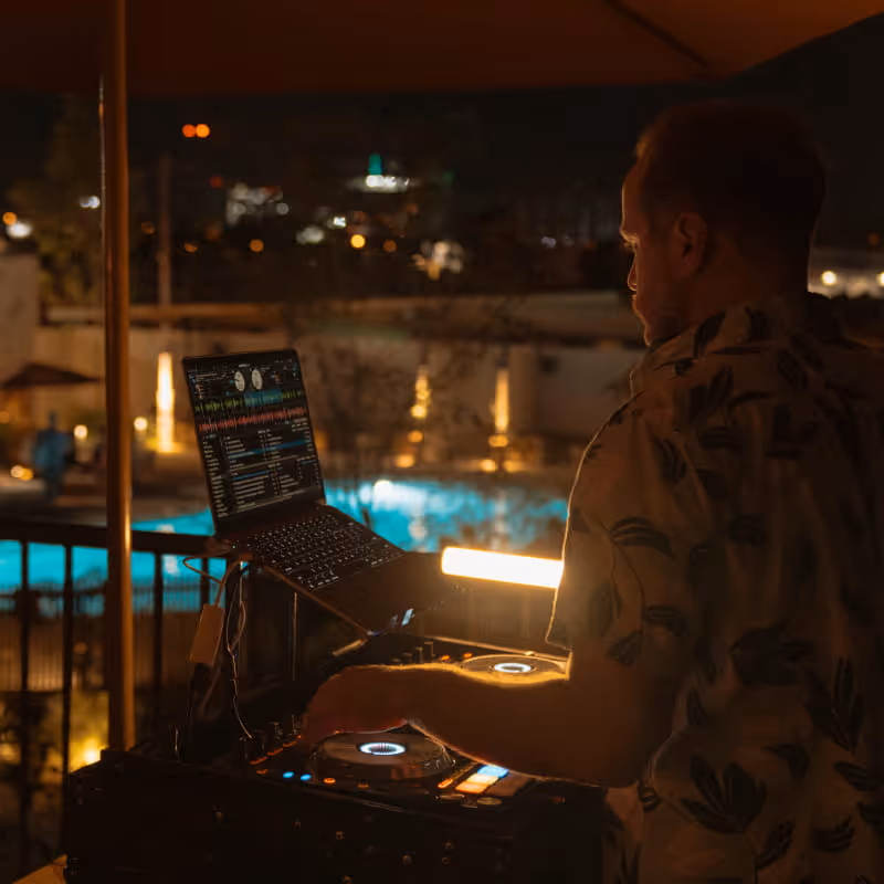 A DJ mixes music on a laptop and turntable setup overlooking a glowing nighttime pool at an outdoor venue.