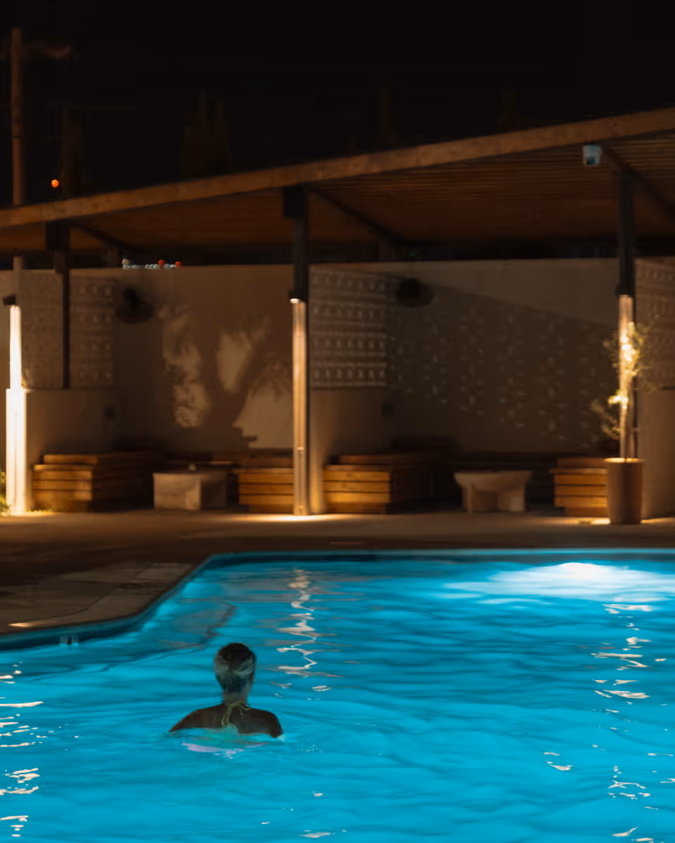 A lone swimmer floats in a glowing turquoise pool at night, surrounded by warm ambient lighting and covered cabanas.