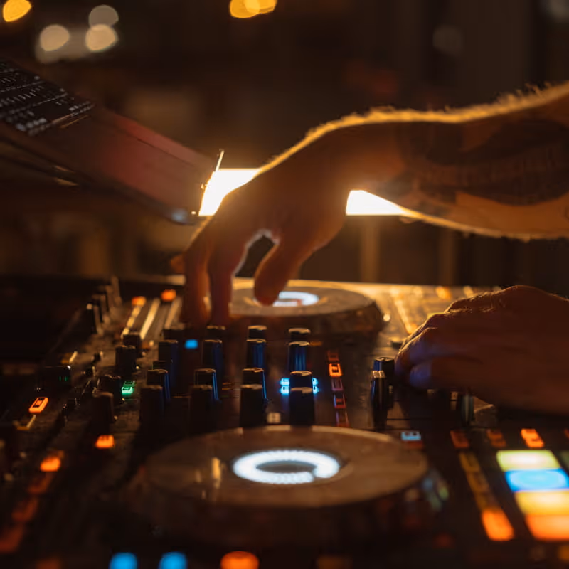 DJ mixing music on an illuminated controller with colorful LED lights and jog wheels in a dark nightclub setting.