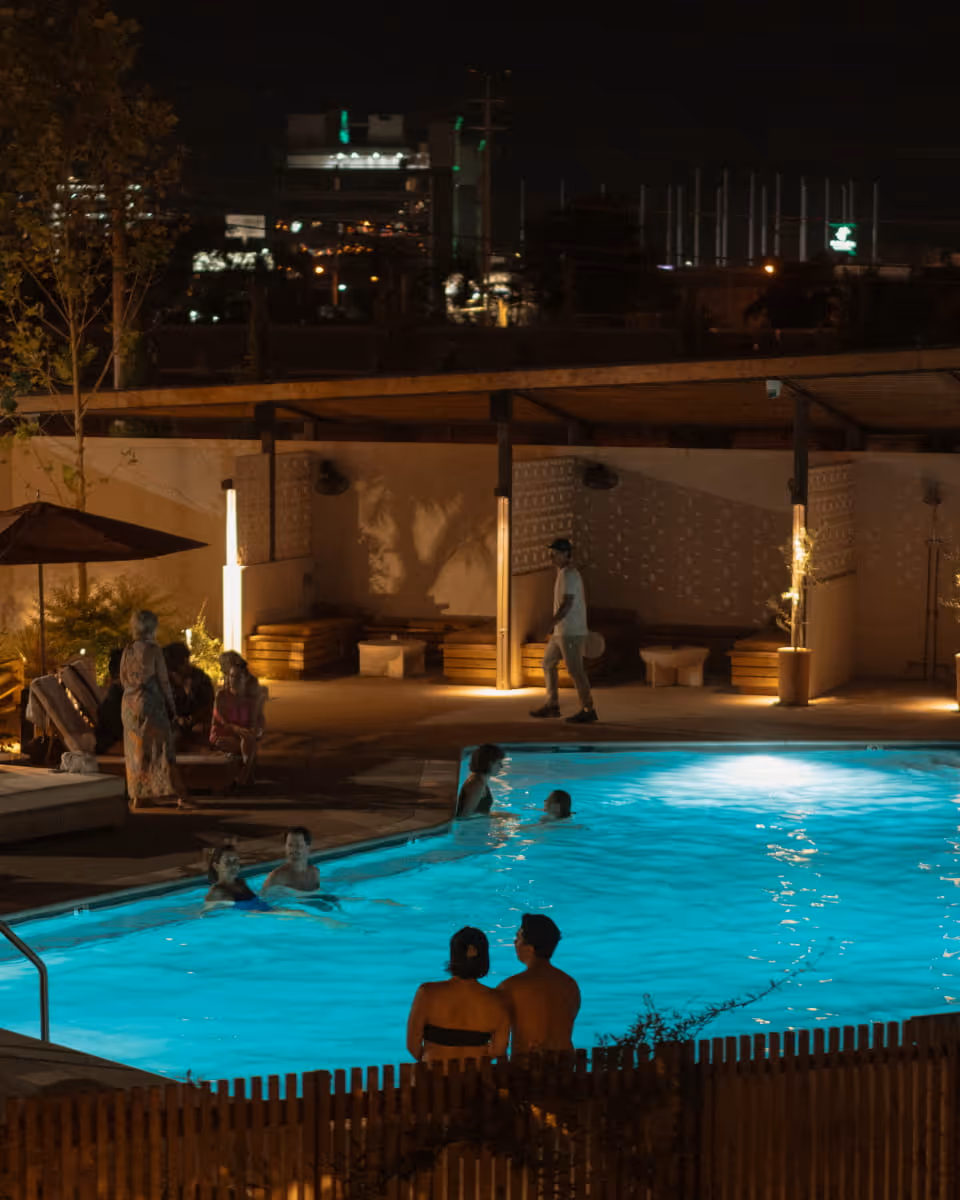 Guests swim and socialize around a brightly lit turquoise pool at night, with city lights visible in the background.