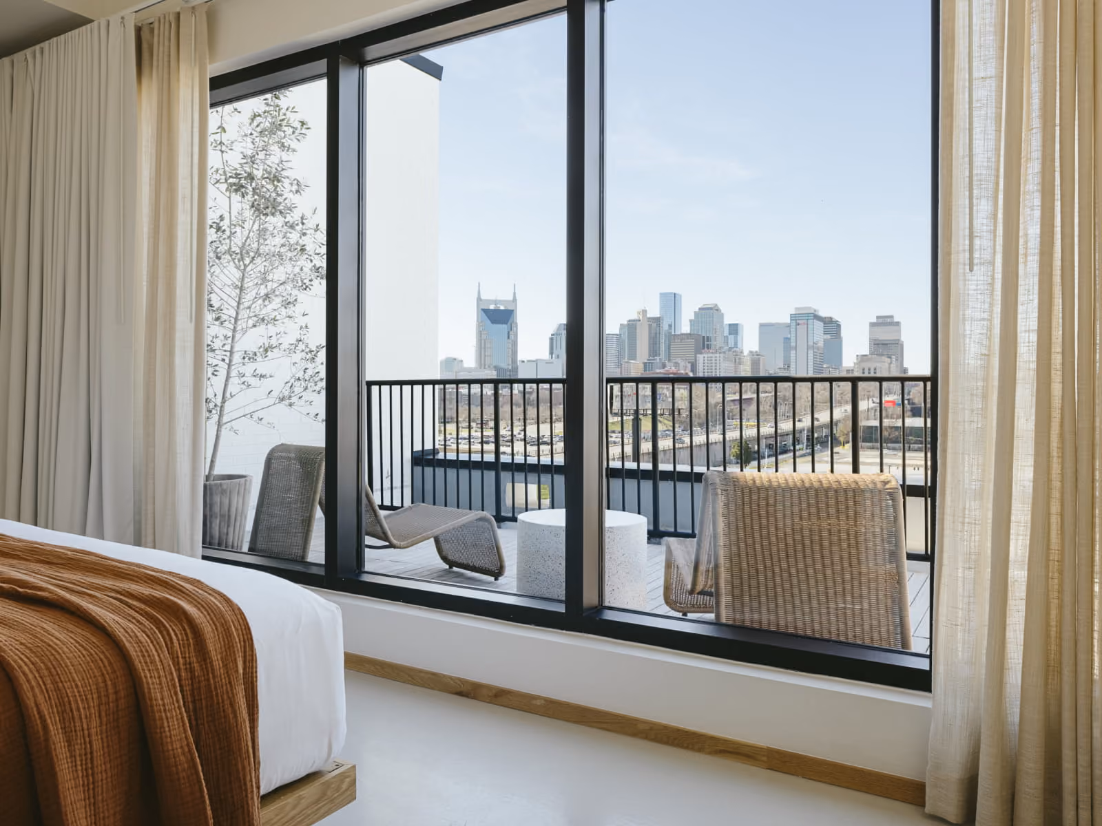 Modern hotel bedroom with rust-colored throw blanket, floor-to-ceiling windows, and balcony overlooking Nashville skyline