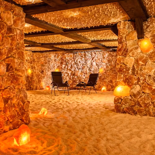 A salt cave therapy room with two lounge chairs, Himalayan salt lamps, salt-covered walls, and a sandy floor.