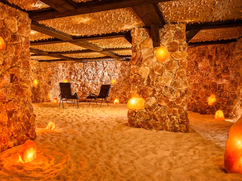 A salt cave therapy room with two lounge chairs, Himalayan salt lamps, salt-covered walls, and a sandy floor.