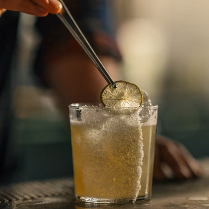 A hand stirs a margarita cocktail garnished with a lime wheel in a salt-rimmed glass on a bar counter.