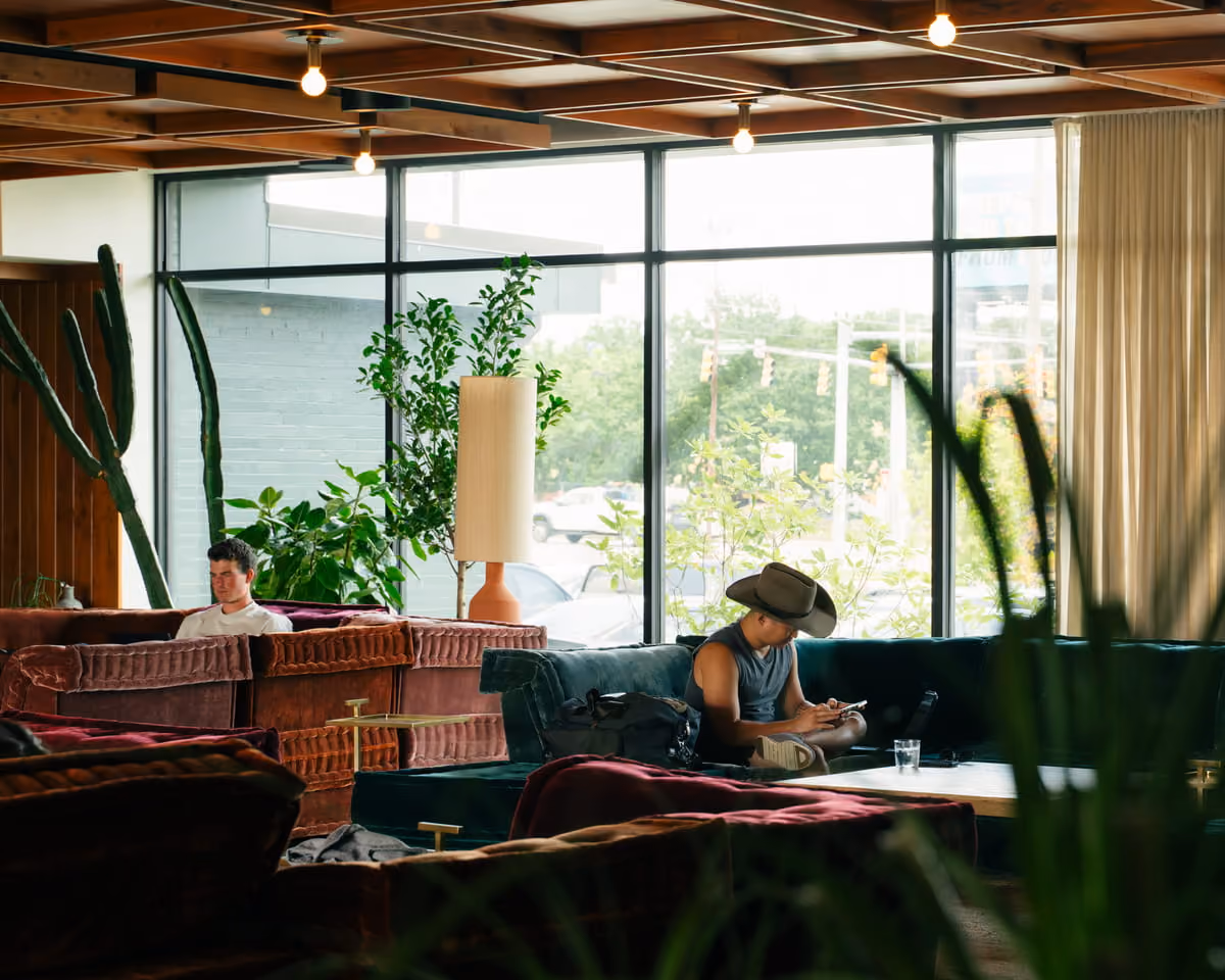 Two people relax on plush velvet couches in a plant-filled hotel lobby with large windows and warm wood ceiling.