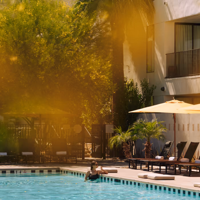 A woman relaxes in a sunlit hotel swimming pool beside lounge chairs and a yellow umbrella on a warm day