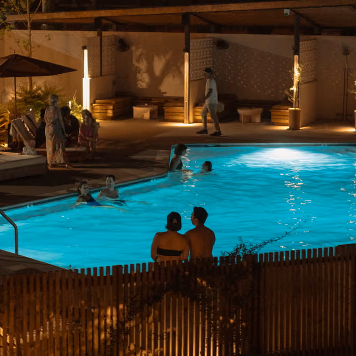 Guests swimming and relaxing by a glowing turquoise pool at night while a couple sits together at the fence
