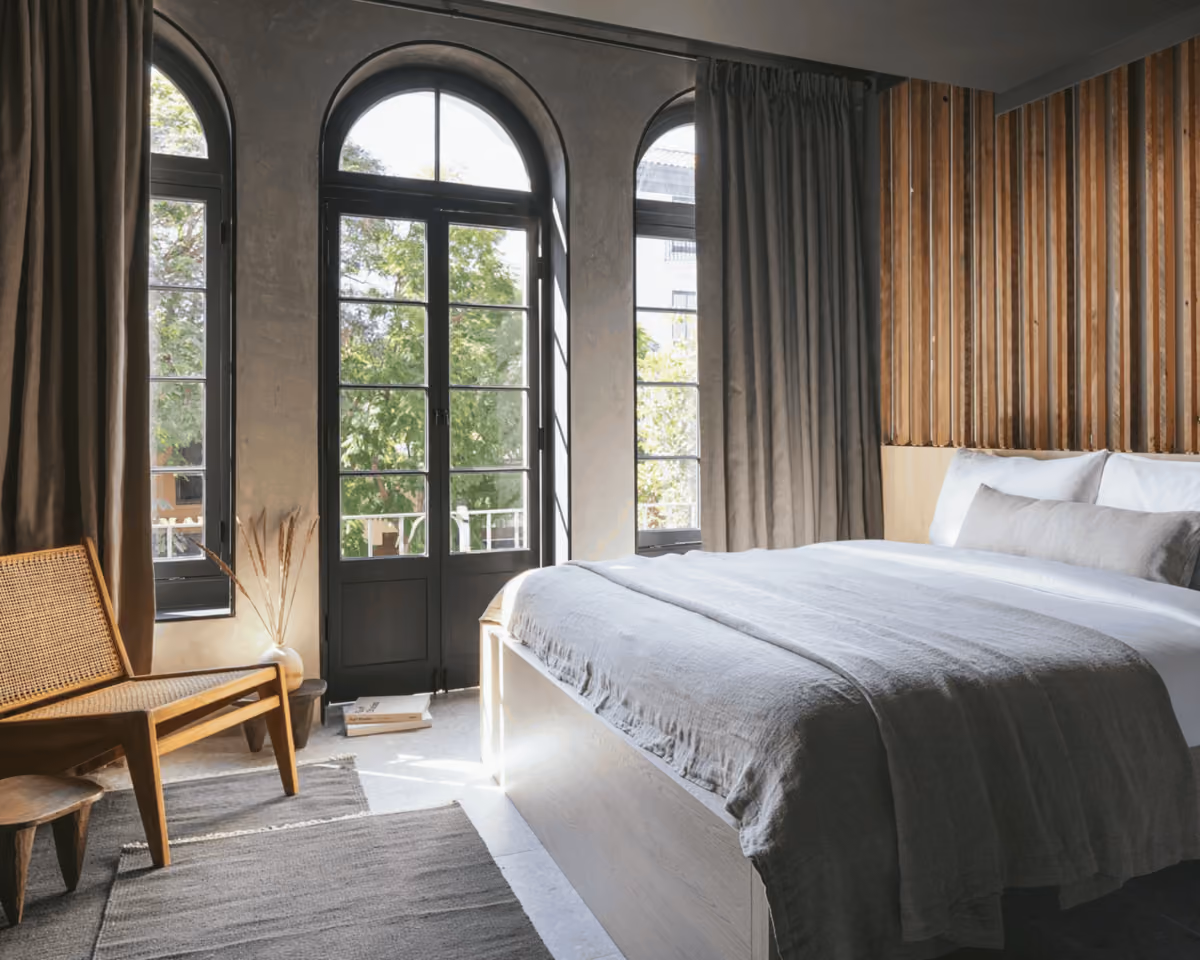 Modern boutique hotel bedroom with arched French doors, wood slat headboard, linen bedding, and mid-century rattan chair