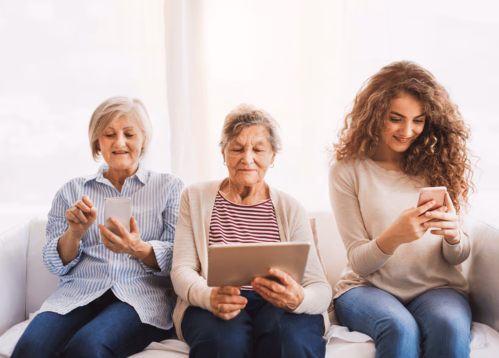 Three generations of women sitting on a couch, each using a tablet or phone and smiling