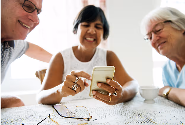 Three elderly people smiling and looking at a smartphone together at a table with glasses and a cup.