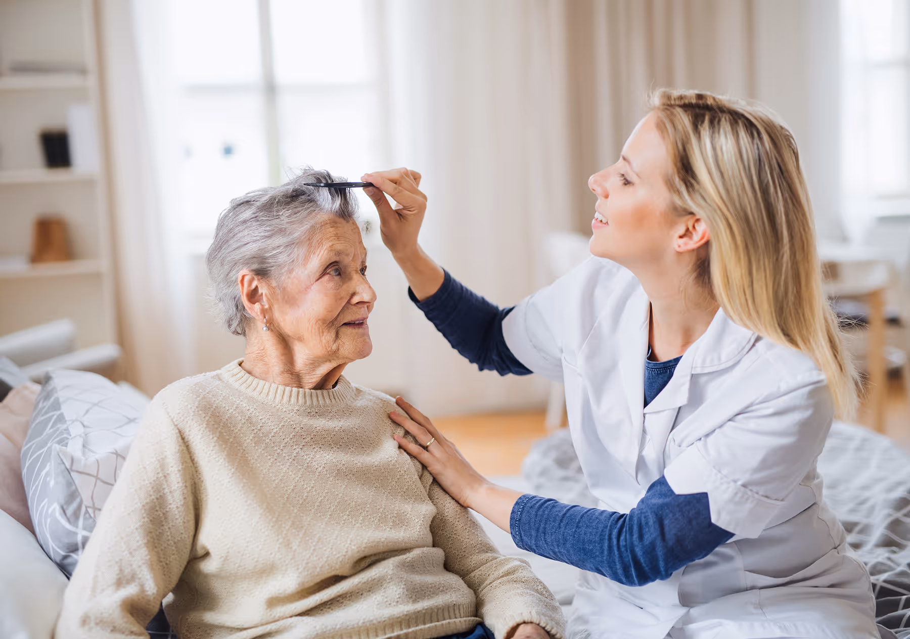 Caregiver combing elderly woman's gray hair while sitting together in a cozy room.