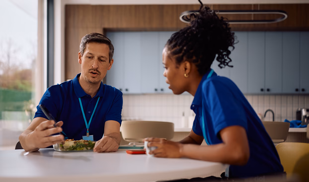 Two people in blue shirts sitting at a table in a modern kitchen, with one person holding a fork over a meal container and the other holding a cup.
