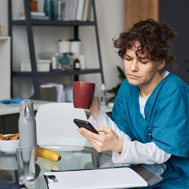 A woman in teal medical scrubs sits at a glass table holding a red mug in one hand and looking at a smartphone in the other, with a clipboard, banana, water bottle, and empty glass also on the table.