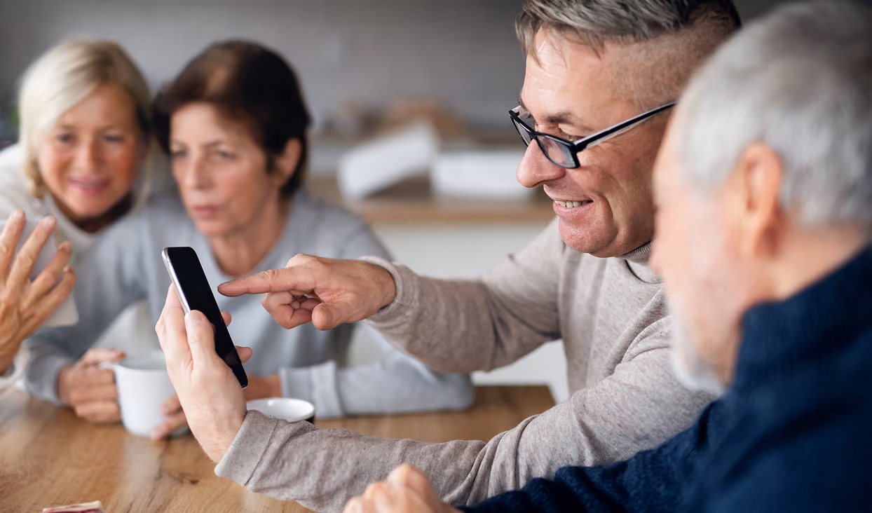 Group of four older adults sitting around a table, smiling and looking at a smartphone held by one man wearing glasses.