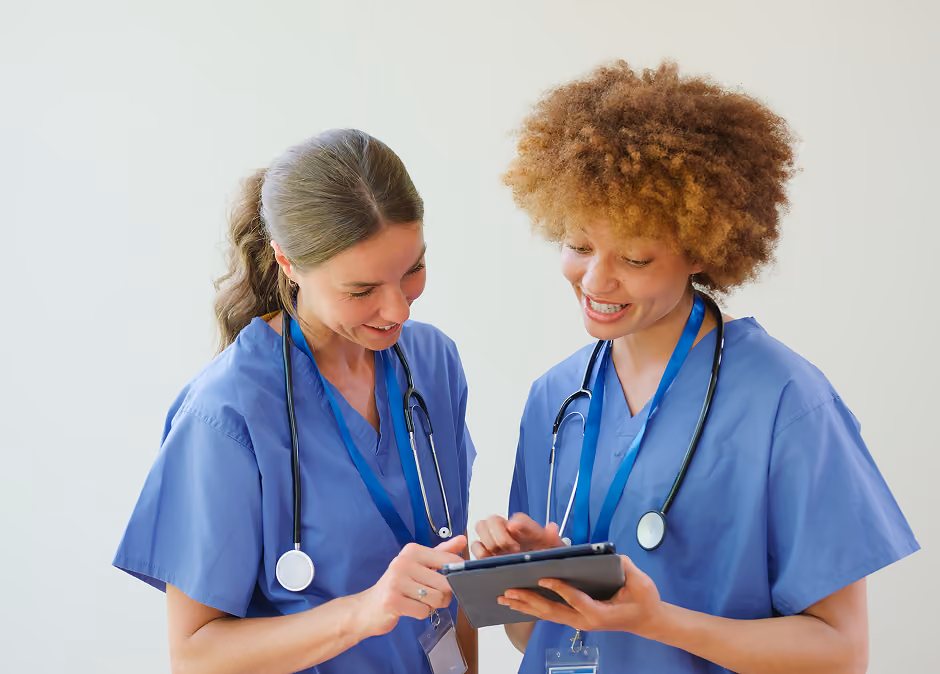 Two female healthcare professionals in blue scrubs with stethoscopes around their necks, smiling and looking at a tablet together against a plain light background.