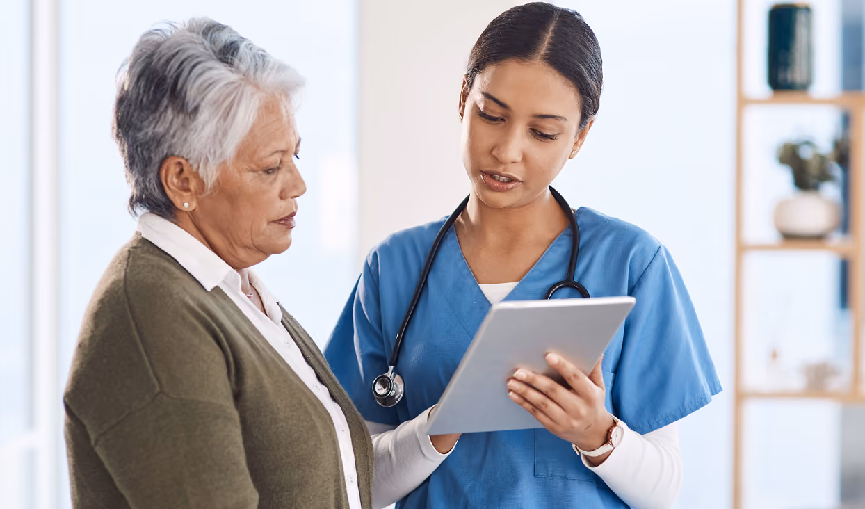 A nurse in blue scrubs with a stethoscope around her neck talks to an elderly woman while showing her information on a digital tablet in a bright medical office.