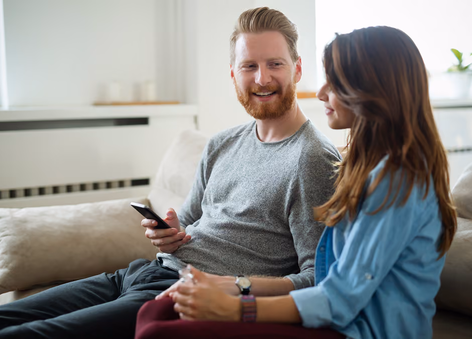 A smiling man holding a smartphone sitting on a couch next to a woman who is looking at him, both appear engaged in conversation in a bright living room.