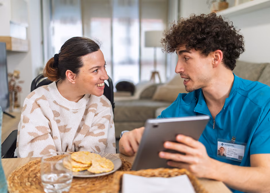 A male healthcare worker in blue scrubs holding a tablet and talking to a smiling woman in a wheelchair at a dining table with plates of food and a glass of water.